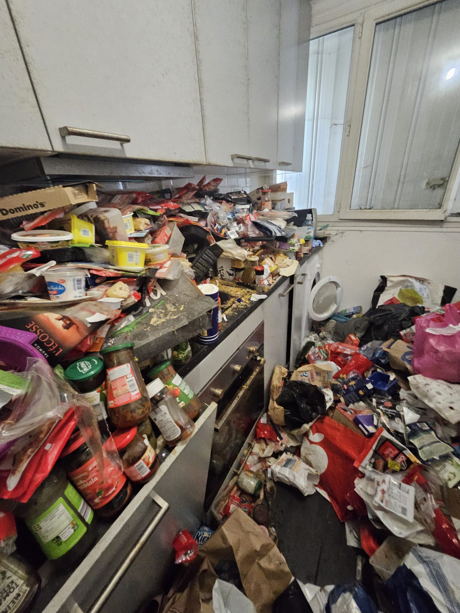Cuisine envahie de cartons Domino's, conserves, bocaux, emballages plastique sur plusieurs niveaux